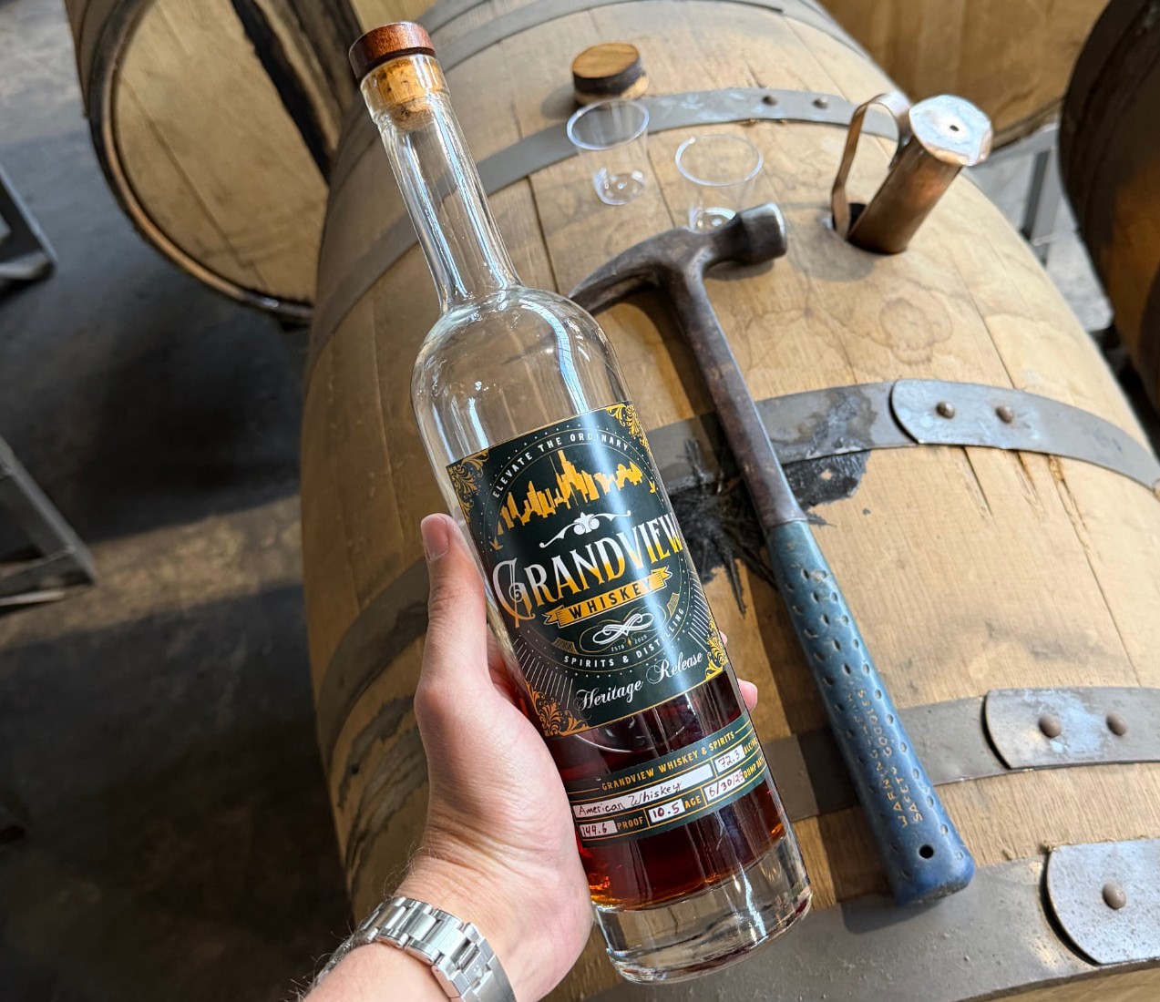 A hand holding a bottle of Grandview whiskey in front of an oak barrel in a warehouse
