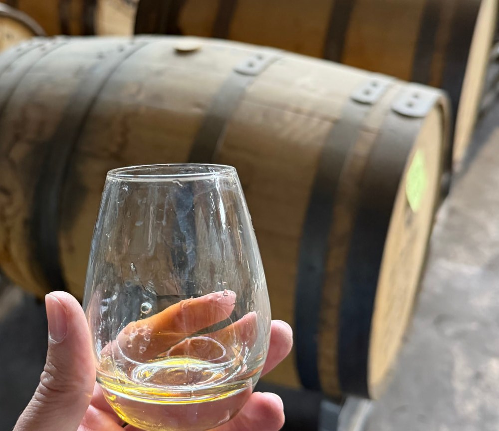 A hand holding a tasting glass of amber bourbon in a barrel warehouse