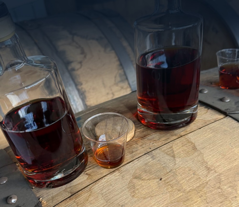 Two glass bottles and tasting glasses resting on an oak barrel in a warehouse